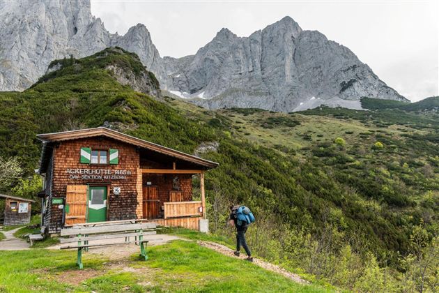 Eine Berghütte inmitten von grünen Wiesen und hohen Bergen. Ein Wanderer nähert sich der Hütte.