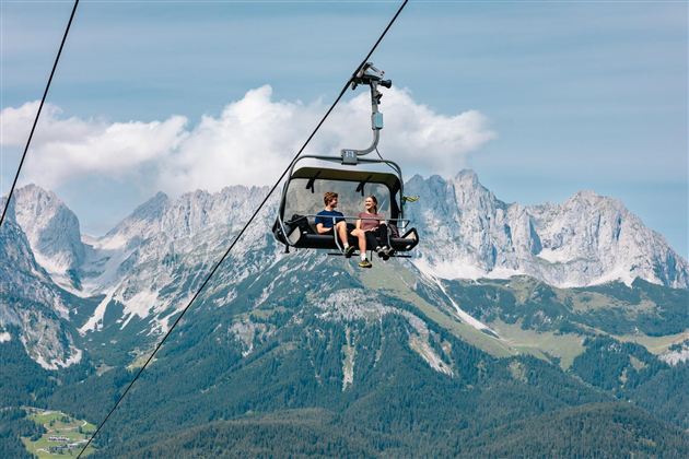 Zwei Personen in einem Sessellift vor dem Bergmassiv des Wilden Kaisers unter blauem Himmel.