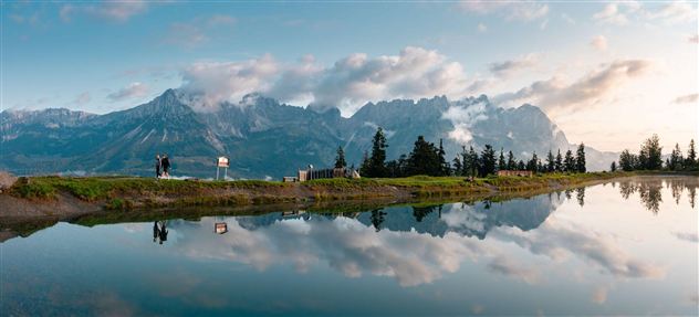 Eine beeindruckende Berglandschaft mit spiegelndem Wasser im Vordergrund. Der Himmel ist teilweise bewölkt, und Bäume umrahmen die Szene.