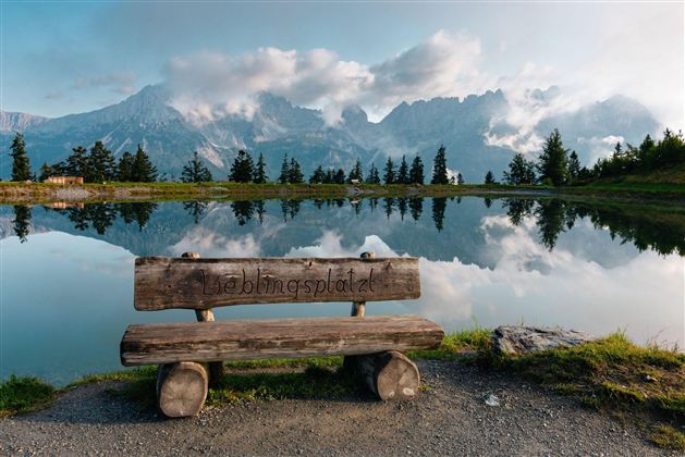 Eine Holzbank steht am Ufer eines ruhigen Sees, umgeben von Bergen. Der Himmel ist teilweise bewölkt und spiegelt sich im Wasser.