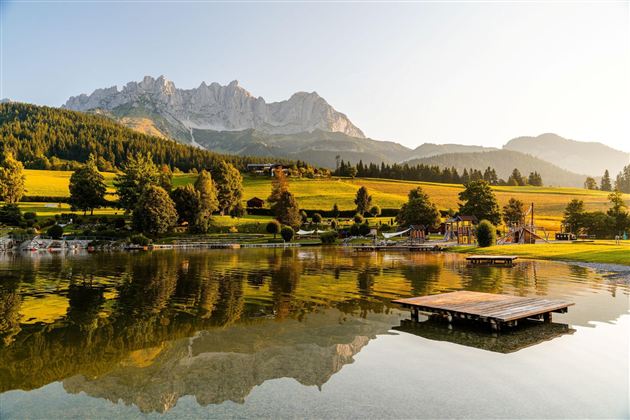 Ein malerischer See mit sanften Hügeln und reflektierenden Bergen im Hintergrund. Die goldene Abendsonne beleuchtet die Landschaft und schafft eine ruhige Atmosphäre.