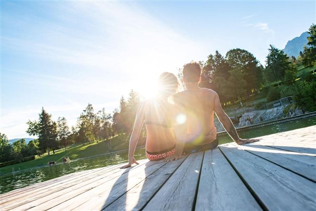 Ein Paar sitzt auf einem Holzsteg am Wasser und genießt den Sonnenuntergang. Die Atmosphäre ist entspannt und romantisch mit einer schönen Naturkulisse.