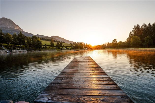 Ein malerischer Steg führt über einen ruhigen See. Die Sonne geht hinter den Bergen auf und taucht die Landschaft in warmes Licht.