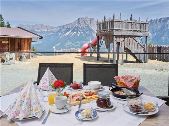 A set table with breakfast outdoors, including pastries, jam, and coffee. In the background, a playground with a slide and the mountains can be seen.