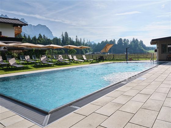 Ein schöner Pool mit klarem Wasser, umgeben von grünen Wiesen und Bergen. Liegen und Sonnenschirme laden zum Entspannen ein.