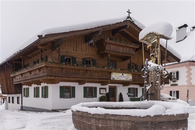 Ein traditionelles Holzhaus im Schnee mit grünen Fensterläden. Im Vordergrund befindet sich ein Brunnen, umgeben von einer winterlichen Landschaft.