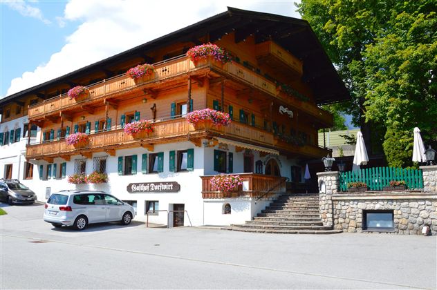 A traditional wooden house with balconies decorated with flowers. Additionally, a green surrounding and a parking lot in front.