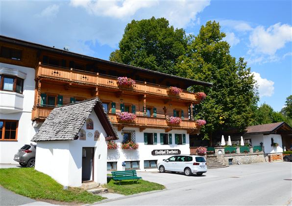 A traditional old building with a wooden balcony and colorful flowers. It is located in a quiet environment with trees and an asphalt road.