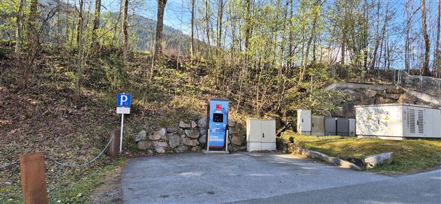 Een parkeerplaats met een automaat nabij een bosrijke heuvel. Op de achtergrond zijn enkele techniekcontainers te zien.