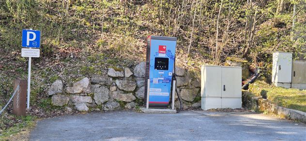 Een parkeerplaats met een automatische ticketautomaat staat op de voorgrond. Op de achtergrond zijn bomen en een rotsachtige helling te zien.