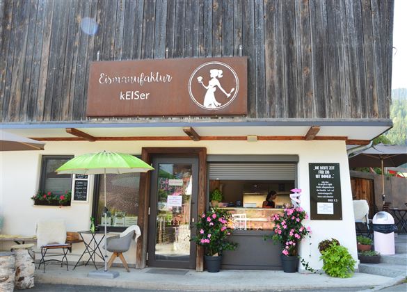 Ein einladendes Geschäft mit der Beschriftung „Ernährungskultur keISer“. Es gibt Pflanzen und Möbel im Außenbereich, darunter einen Sonnenschirm.