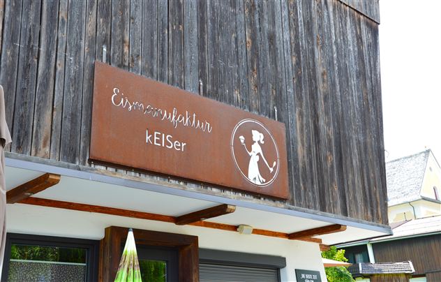 Ein Holzschild mit der Aufschrift "Eismanufaktur kElSer". Im Hintergrund ist ein modernes Gebäude mit einem klaren Himmel zu sehen.