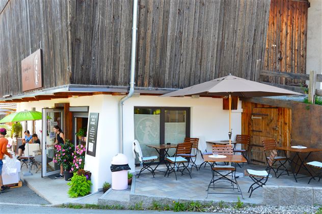 Ein gemütliches Café mit Holzverkleidung und Tischen im Freien. Es gibt Sonnenschirme und Blumen vor dem Eingang.