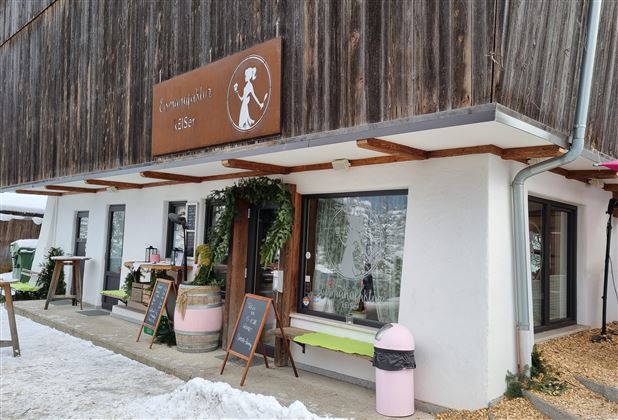 Ein gemütliches Café mit einer rustikalen Holzfassade und großen Fenstern. Der Eingangsbereich ist einladend mit Pflanzen und Tischen im Freien.