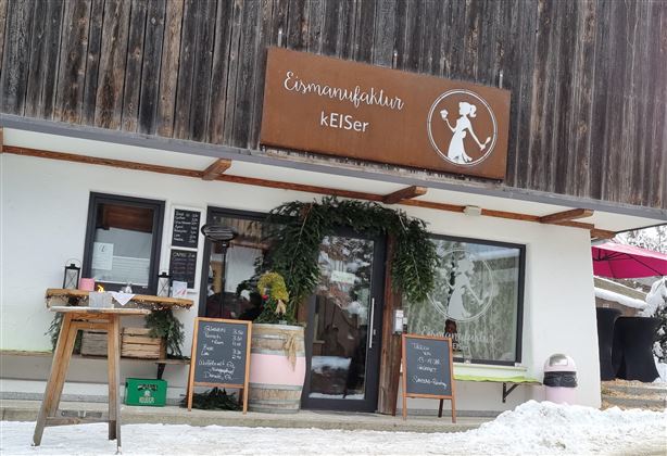 Ein gemütliches Café mit einem rustikalen Holzschild. Draußen liegen Schnee und dekorative Pflanzen.
