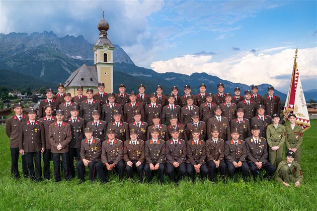 Eine Gruppe von Männern und Frauen in Uniform steht in einer grünen Wiese vor einer Kirche und Bergen im Hintergrund. Sie posieren gemeinsam für ein Gruppenfoto.