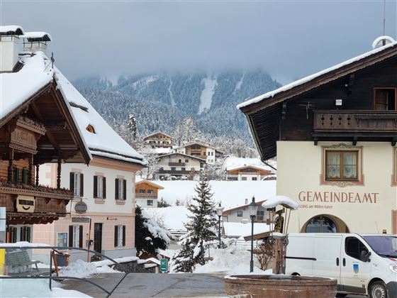 Eine malerische Winterlandschaft mit schneebedeckten Dächern und Bergen im Hintergrund. Im Vordergrund sind traditionelle Gebäude und ein Fahrzeug zu sehen.