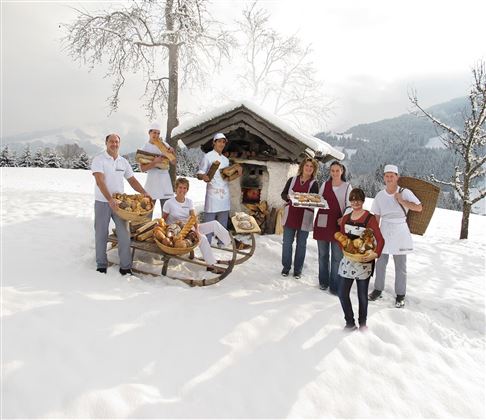 Eine Gruppe von Menschen steht im Schnee vor einer kleinen Hütte. Sie halten frisches Brot und Gebäck in den Händen.