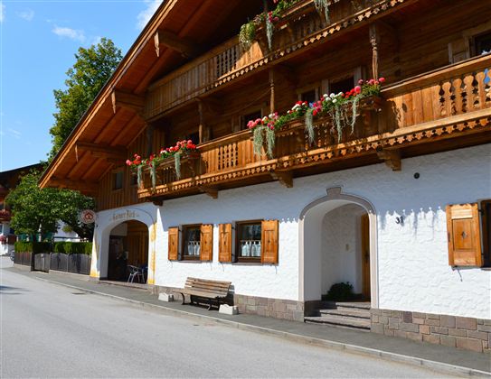 A traditional chalet with wooden balustrades and flower boxes. The street in front of the building is quiet and lined with trees.