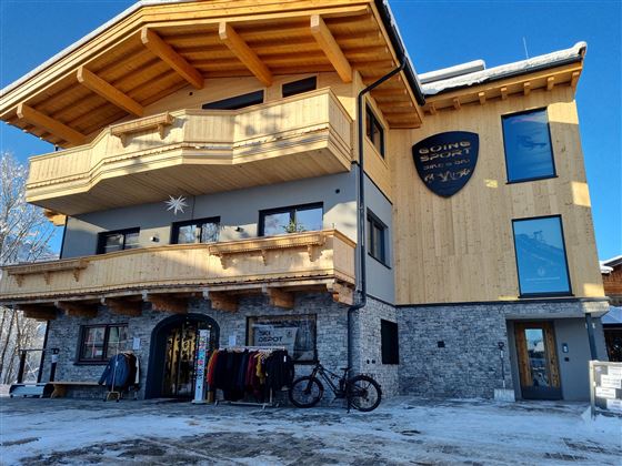 A modern building in alpine style with a wood and stone facade. In front of the house, clothing is for sale and a bicycle.
