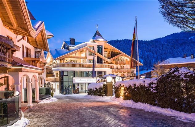 Ein malerisches Alpenhotel im Winter bei Dämmerung. Der Schnee bedeckt den Weg und die umliegenden Bäume, während die Lichter des Gebäudes warm leuchten.