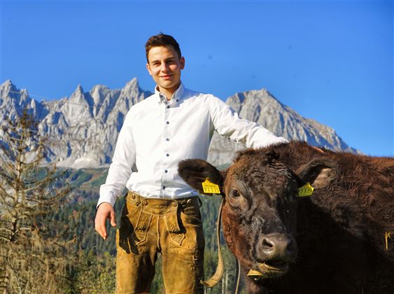 Ein junger Mann steht neben einem schwarzen Rind in einer Berglandschaft. Im Hintergrund sind beeindruckende Berge und ein blauer Himmel zu sehen.