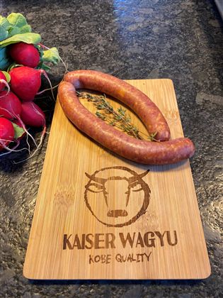 Eine Holzplatte mit der Aufschrift "Kaiser Wagyu" zeigt zwei Würste und frische Radieschen. Die Würste liegen auf einem Küchentisch und wirken appetitlich.