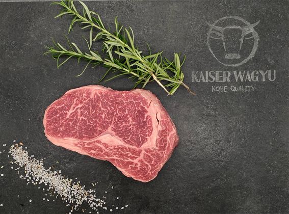 Ein hochwertiges Stück Wagyu-Rindfleisch liegt auf einem dunklen Untergrund. Daneben sind frische Kräuter und Salzstreusel zu sehen.