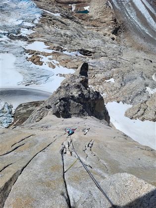 Ein Kletterer erklimmt eine steile Felswand mit Blick auf eine beeindruckende Berglandschaft. Schnee und Gletscher sind in der Umgebung sichtbar.