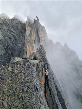 Ein Kletterer bestigt einen steilen, felsigen Gipfel mit Nebel in der Umgebung. Die dramatische Landschaft zeigt hohe Felsen und eine mystische Atmosphäre.