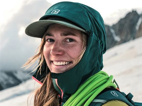 Eine lächelnde Person mit einem grünen Hut und wetterfester Kleidung steht im Gebirge. Im Hintergrund sind Wolken und Berglandschaften zu sehen.