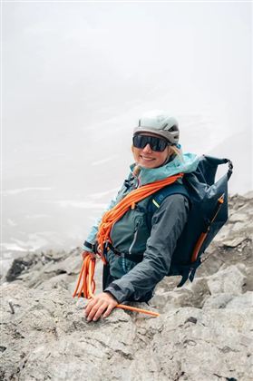 Eine Kletterin steht auf einem Felsen und trägt einen Helm sowie eine Ausrüstung. Sie lächelt in die Kamera und wirkt bereit für das Abenteuer in der Berglandschaft.