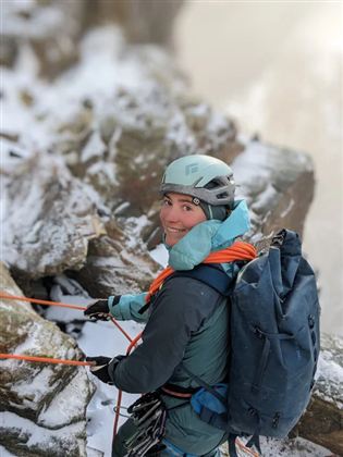 Eine Person beim Klettern in schneebedecktem Gelände. Sie trägt einen Helm und Rucksack und hält Seile in der Hand.