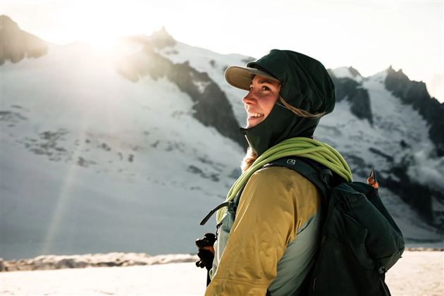 Eine Person steht am Berg und schaut lächelnd in die Ferne. Die Landschaft ist schneebedeckt und die Sonne scheint hell.