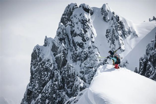 Ein Skifahrer steht auf einem schneebedeckten Gipfel mit beeindruckenden Felsen im Hintergrund. Die Szene vermittelt ein Gefühl von Abenteuer in einer winterlichen Berglandschaft.