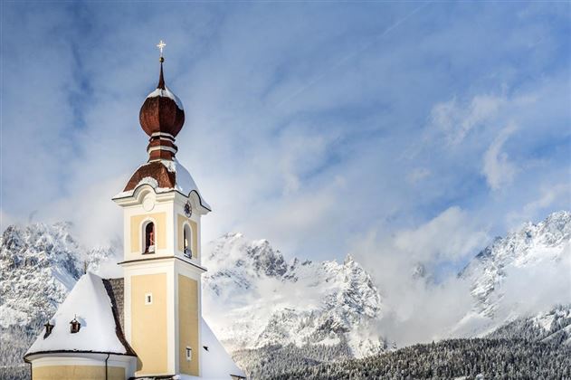 Een schilderachtige kerk met een kenmerkende ui-vormige torenspits, omringd door besneeuwde bergen. De lucht is helder en het landschap lijkt vredig en winters.