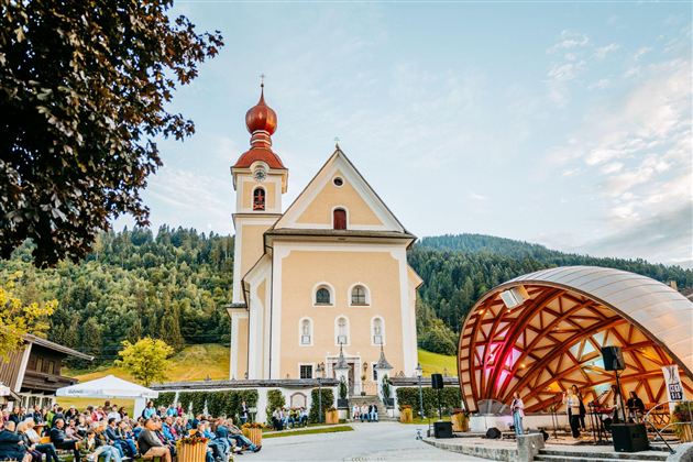 A picturesque church with a red tower stands in the background. In front of the church, a concert is taking place in a wooden pavilion, surrounded by an audience and green nature.