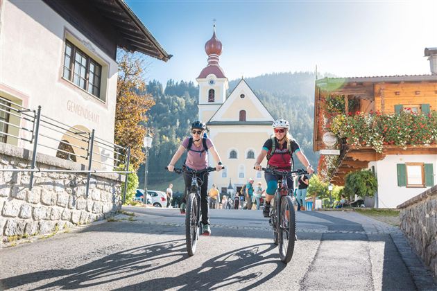 Two women are riding their bicycles through a picturesque village. In the background, there is a church, and the surroundings are bordered by mountains.