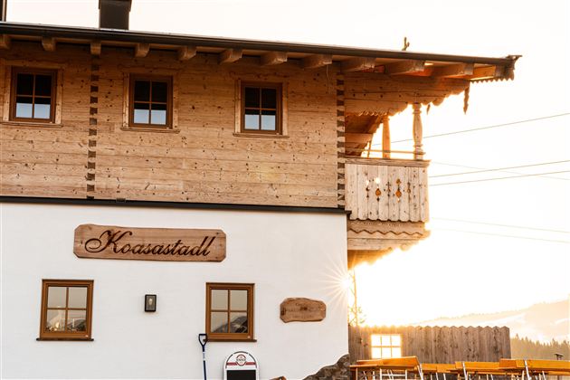 Ein holzverkleidetes Gebäude mit dem Namen "Koaastall". Im Hintergrund ist die Abendsonne zu sehen, die für eine warme Atmosphäre sorgt.