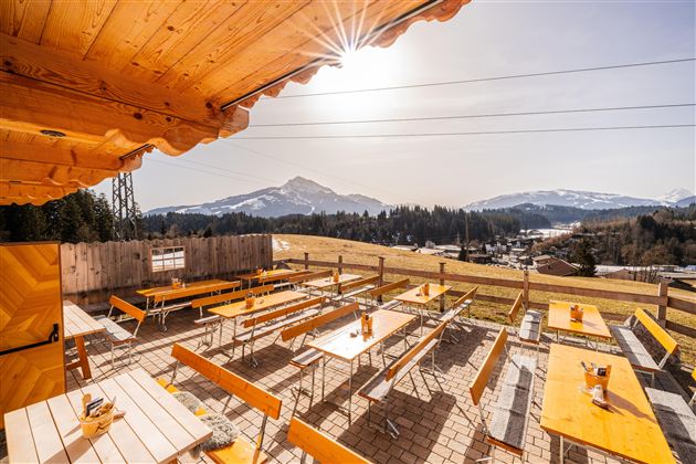 Eine sonnige Terrasse mit Holztischen und Bänken bietet einen Blick auf die Berge. Im Hintergrund ist eine klare, blaue Himmelslandschaft zu sehen.