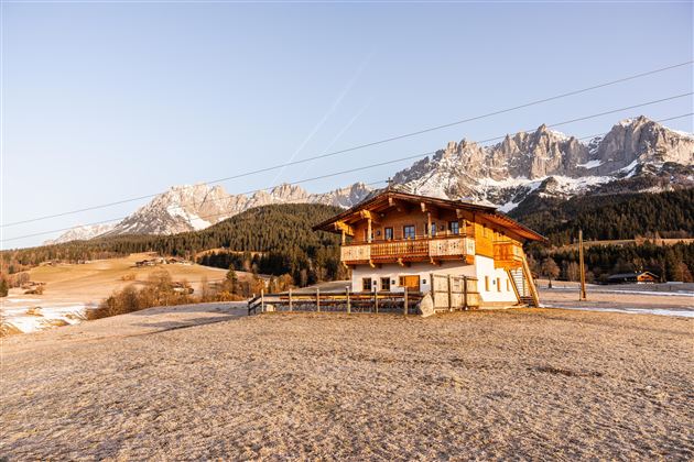 Ein schönes Holzhaus steht in einer winterlichen Landschaft mit schneebedeckten Bergen im Hintergrund. Die Sonne scheint und schafft eine friedliche Atmosphäre.