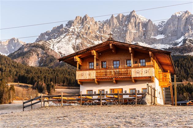 Ein traditionelles Holzhaus in den Bergen, umgeben von schneebedeckten Gipfeln. Die Landschaft ist friedlich und idyllisch.