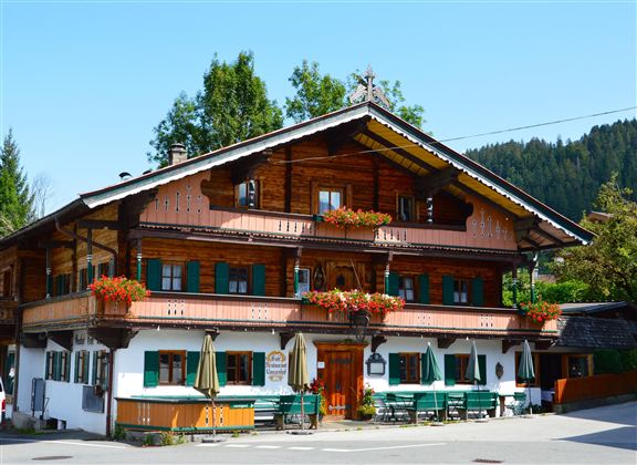 A traditional wooden house in alpine style with green shutters and blooming balconies. It is located surrounded by trees and a clear blue sky.