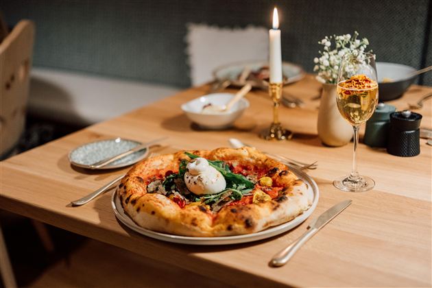 Een tafel met een heerlijke pizza, belegd met verse groenten en een ei. Daarnaast staat een glas champagne en een kaars zorgt voor een gezellige sfeer.
