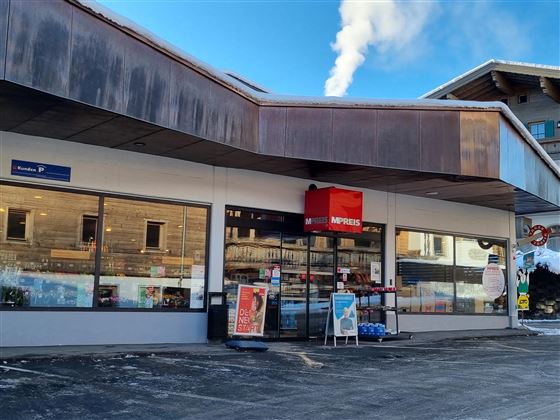Een open winkel met grote ramen en een rood bord. Op de achtergrond zijn enkele gebouwen en een blauwe lucht te zien.