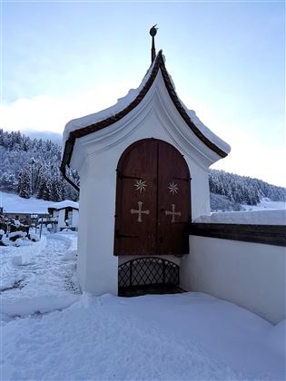Eine schneebedeckte Landschaft mit einem kleinen, weißen Gebäude und einer hölzernen Tür. Im Hintergrund sind schneebedeckte Bäume und ein klarer Himmel zu sehen.