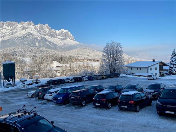 Een parkeerplaats met meerdere auto's en een besneeuwd landschap op de achtergrond. Op de achtergrond zijn bergen en een heldere blauwe lucht te zien.