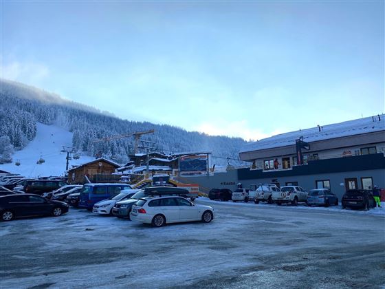 Een parkeerplaats in de bergen, omringd door met sneeuw bedekte heuvels. Op de achtergrond zijn skiliften en gebouwen te zien.