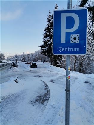 Ein Parkplatzschild mit der Aufschrift "Zentrum" steht in einer verschneiten Umgebung. Im Hintergrund sind schneebedeckte Bäume und ein Weg zu sehen.