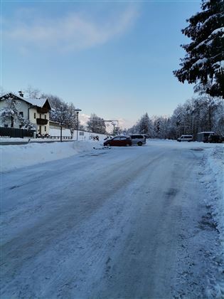 Eine verschneite Straße mit Häusern und Bäumen im Hintergrund. Der Himmel ist klar und blau.